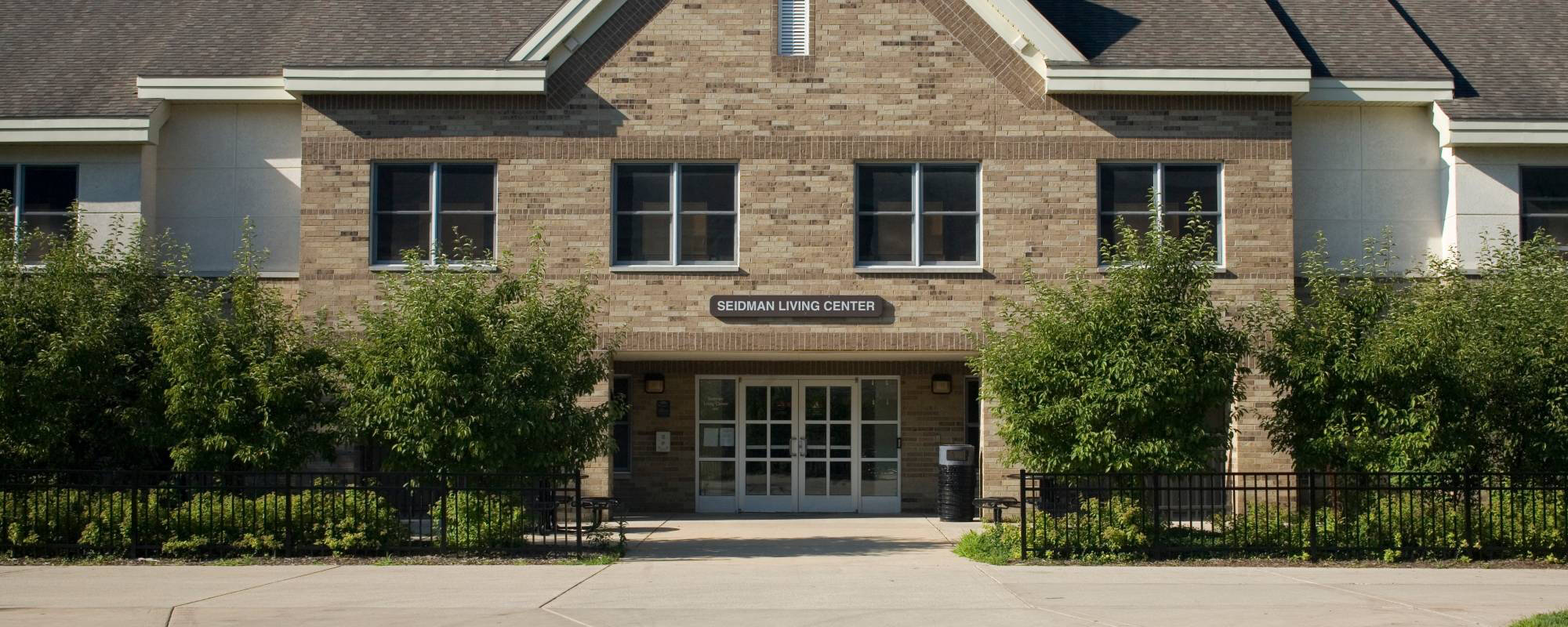 Two-story brick building labeled "Seidman Living Center," featuring several windows, a central entrance, and lush greenery in front, conveying a welcoming atmosphere.